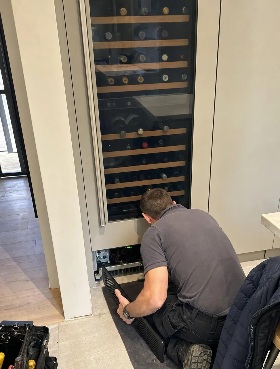 A tall glass-fronted wine column set into a run of pale cabinetry, a technician crouched at its base with the grille removed.