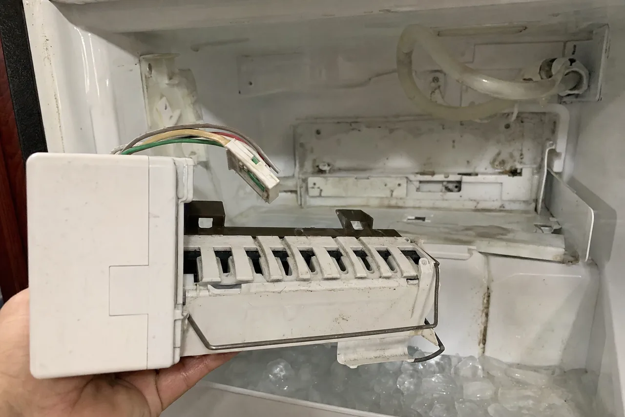 An ice-maker module lifted out on an open palm in front of the freezer compartment it was removed from.