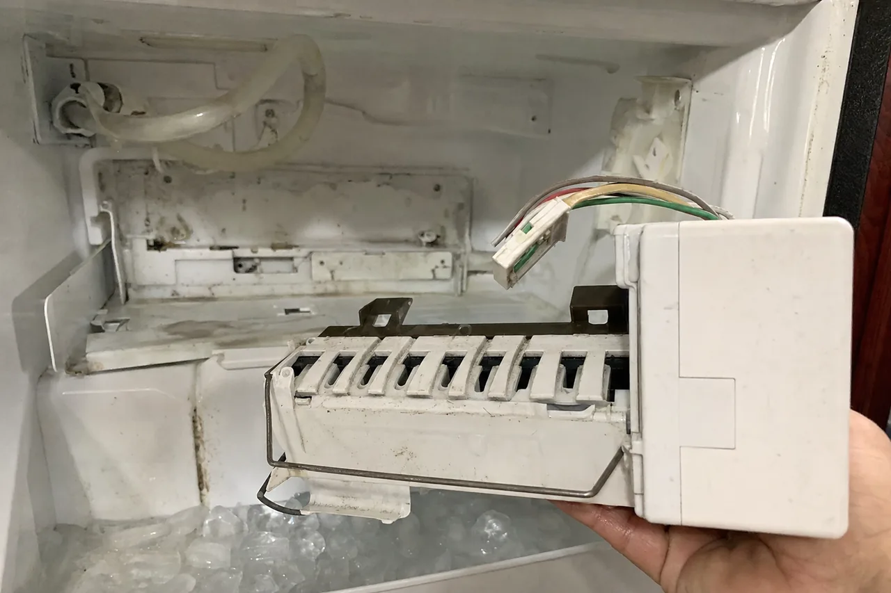 An ice-maker module held out of the freezer on a flat palm, cubes heaped in the bin below the empty bracket.