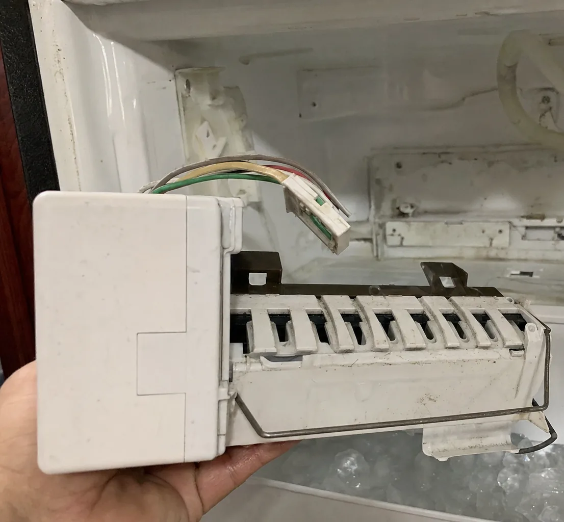 A close view of an ice-maker module in a hand, the mold and ejector arm clearly visible against the freezer wall.