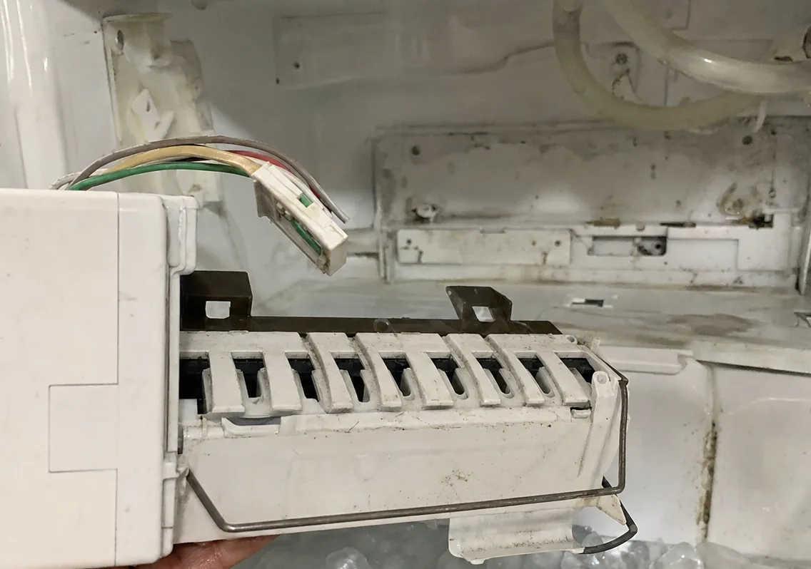 An ice-maker module resting in a hand in front of the empty bracket it came out of, cubes visible in the bin below.