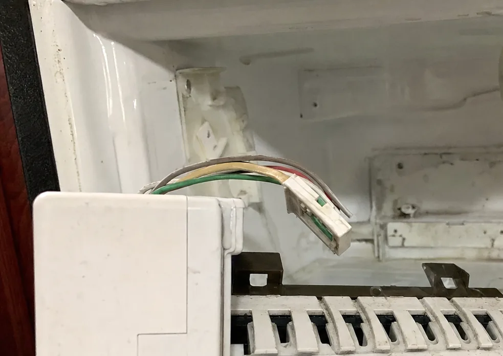 An ice-maker module held to the left of frame with its wiring loom curling back into the freezer liner behind it.