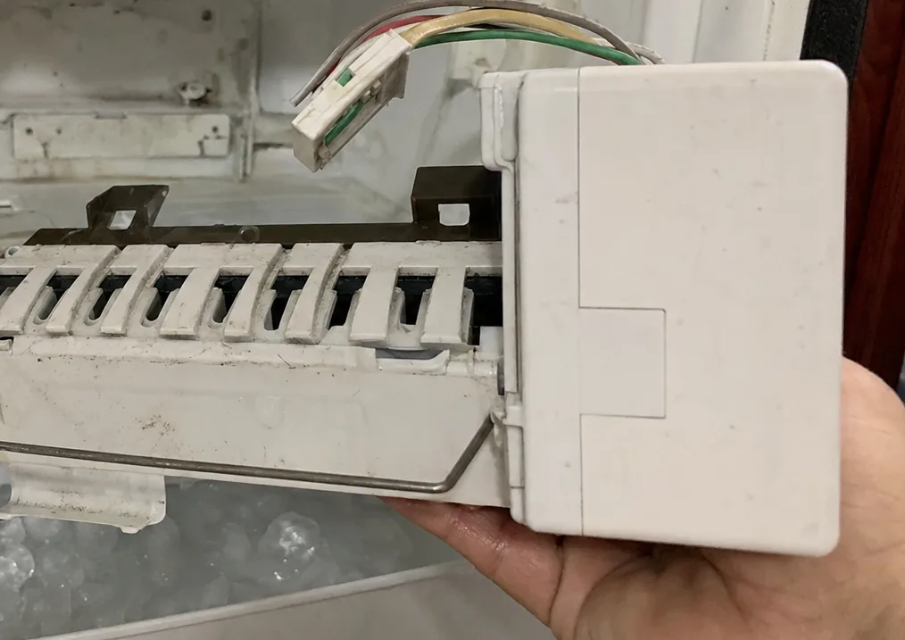 An ice-maker module in a hand at the right of frame, the empty mounting bracket and wiring on the left.