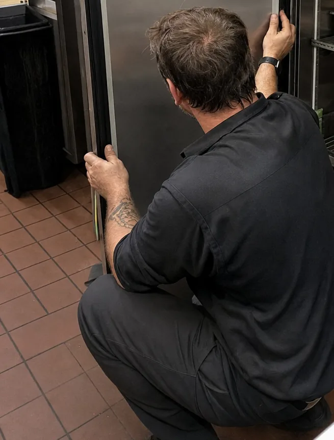 A technician seen from behind, gripping the handle of a refrigerator door on a tiled kitchen floor.